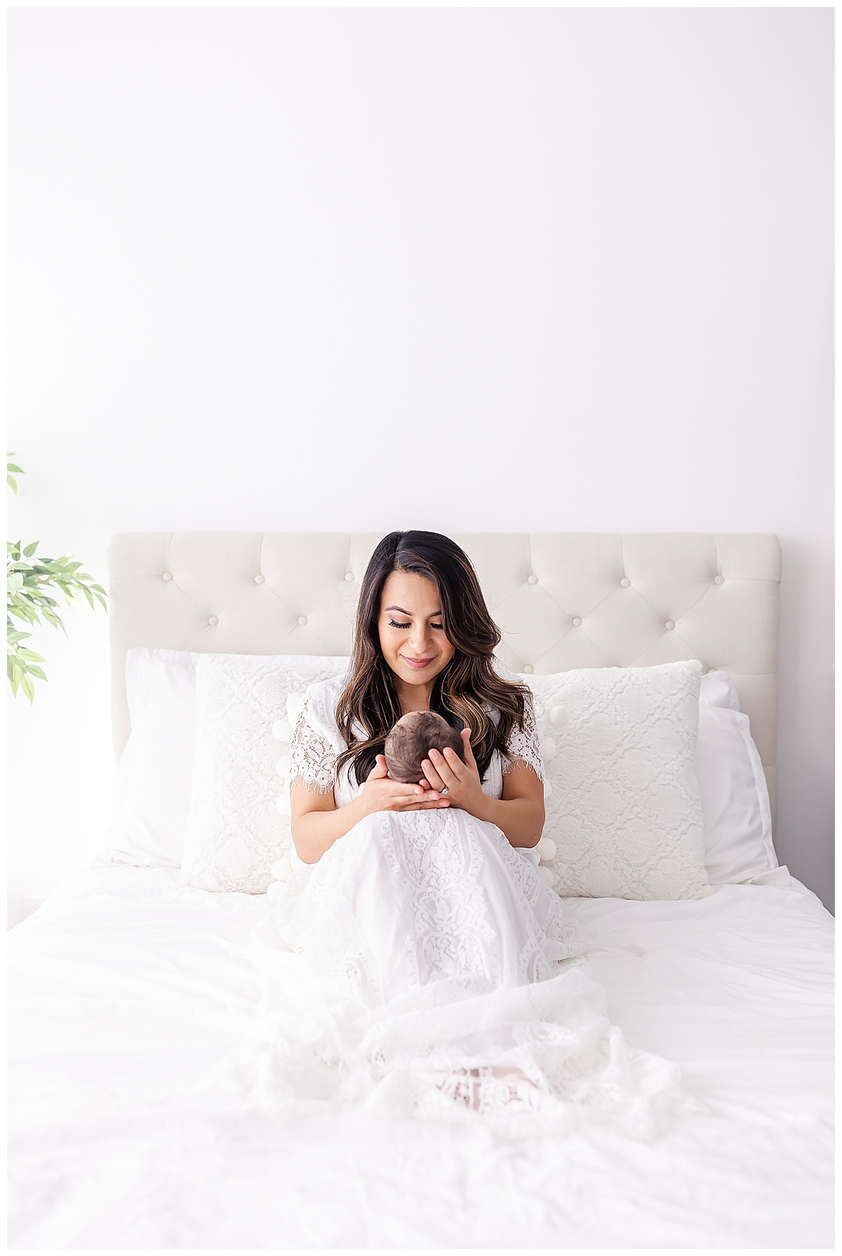 Newborn swaddled in white blanket laying on white bed with mom in white lace dress