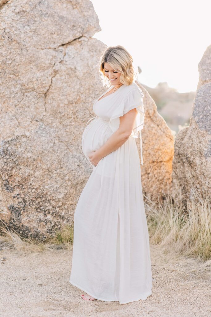 Pregnant woman in cream bohemian dress embraces her belly in a side profile as she smiles at the promise of their future.