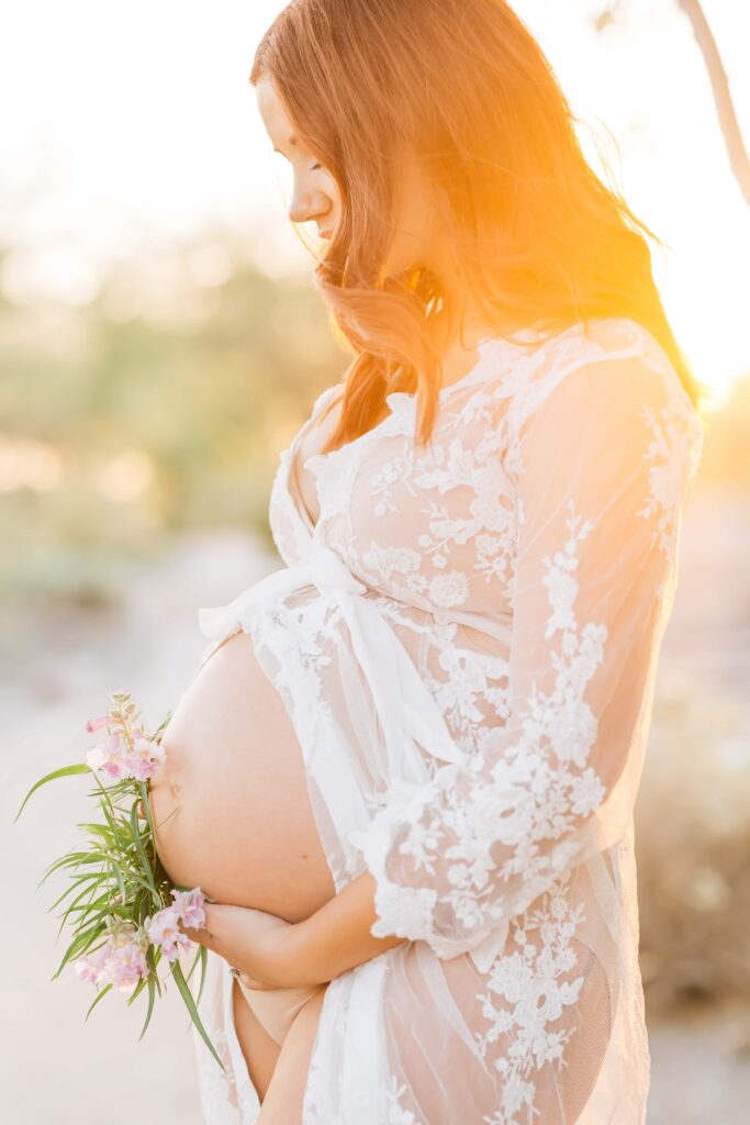 Pregnant mom in white lace robe is photographed in the desert holding flowers. 