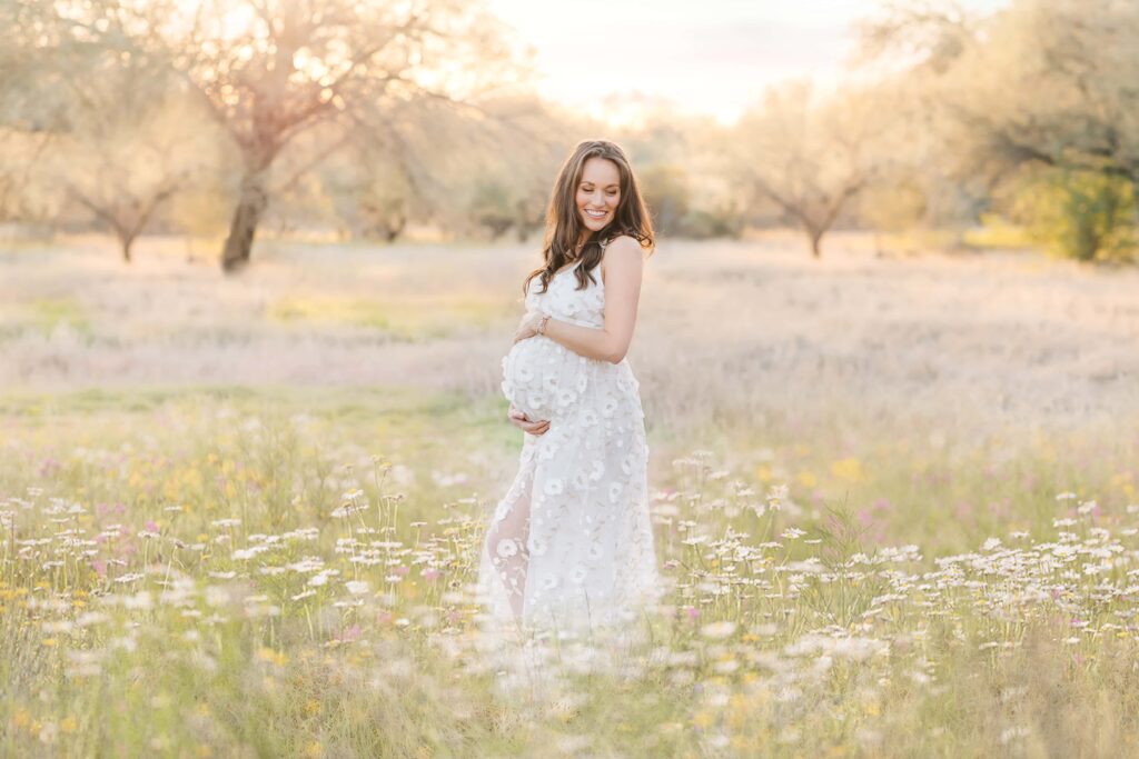 Pregnant woman stands in field of wildflowers while she wraps her arms around her belly. 