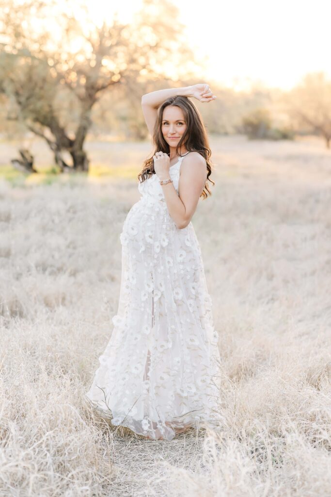 Pregnant woman in cream colored dress with embroidered flowers stands in the dry grass of the Arizona desert. 