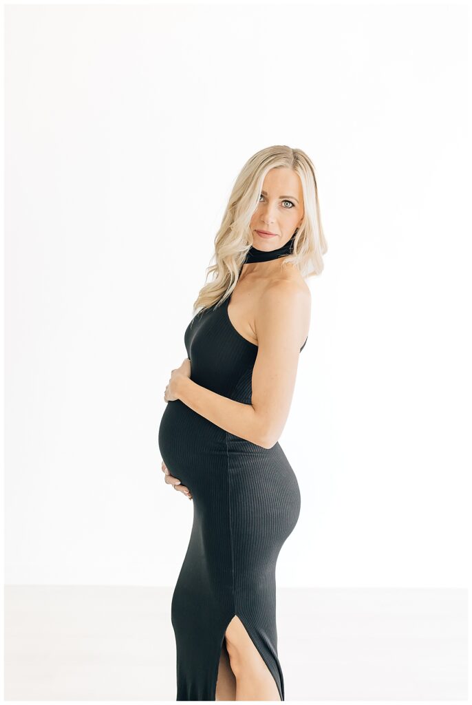 Blonde woman turned to the side, hands on top and bottom of her pregnant belly wearing a formfitting black maternity gown looking directly into the camera on a studio white backdrop 