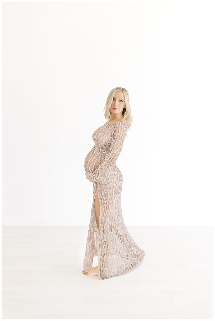 Blonde woman facing camera left, looking directly into the camera, hands on bottom of her pregnant belly wearing a stunning beaded translucent golden hue gown and photographed on a studio white backdrop for her editorial maternity shoot