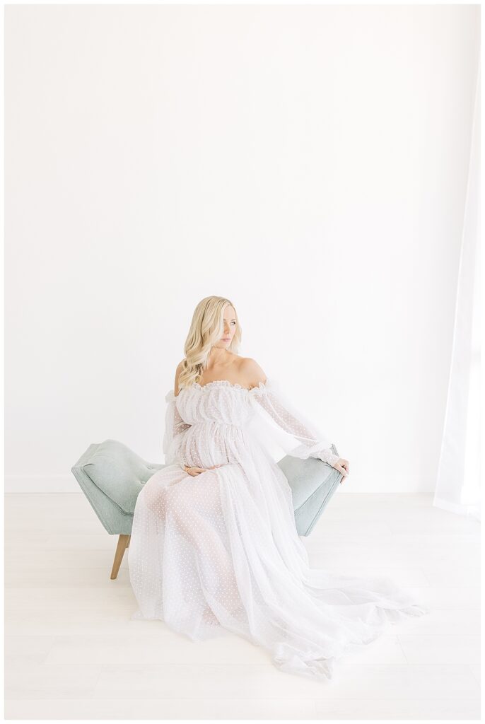 Blonde woman looking off camera right, hands on bottom of her pregnant belly wearing translucent, off the shoulder white dotted, long sleeve maternity gown sitting on a soft sea foam lounge and photographed on a studio white backdrop for her editorial maternity shoot
