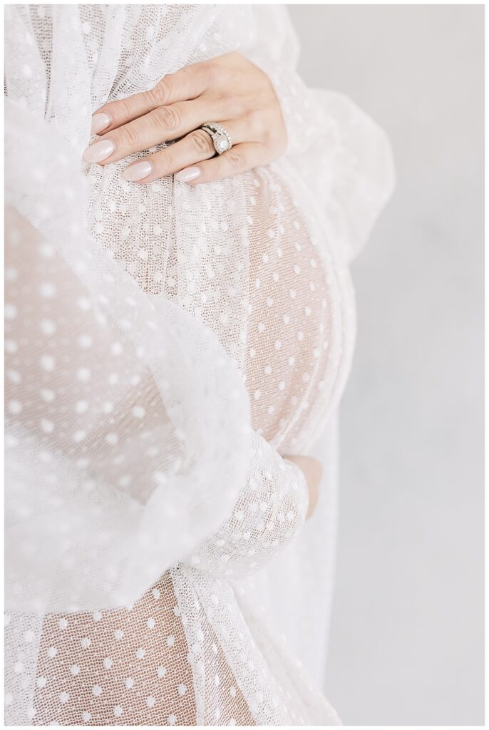 A close up of a mom's pregnant belly from the side, hands on top and bottom of her baby bump wearing translucent, off the shoulder white dotted, long sleeve maternity gown photographed on a studio white backdrop