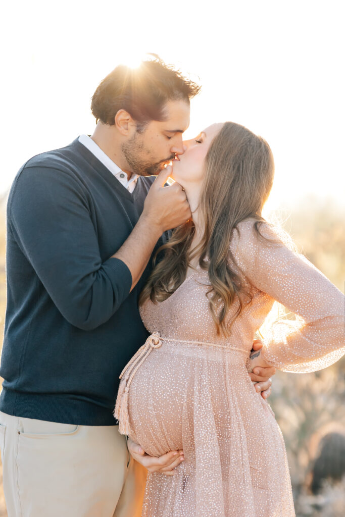 A brunette pregnant woman dressed in a peachy neutral toned long sleeve shimmery dress (camera right)  about to kiss her husband wearing a navy blue sweater overtop a white colored shirt and light tan chinos (camera left) photographed outdoors for a sunset maternity session