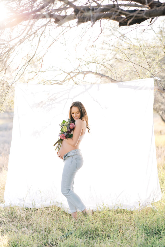 A brunette pregnant woman photographed outdoors in a field with a white sheet hung up in the trees. She is holding her bare belly in one hand and a bright and colorful bouquet in the other wearing light-wash jeans and looking over her shoulder to the ground for this amazing maternity photoshoot idea