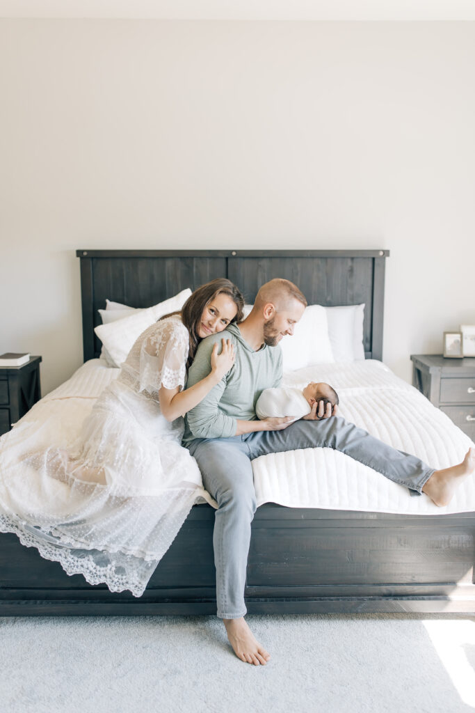 An in-home newborn photography session with a brunette new mom dressed in a white, lace, flowing short-sleeve maternity gown (left) wraps her arms around her husband as she sits on the end of the black wooden master bed.  Dad, sitting side-ways and holding their dark haired swaddled newborn is dressed in a soft green thin sweatshirt and light-wash jeans with bare feet. The room is very neutral cool tones with lots of white and with two identical black wooden nightstands on either side of the bed. 