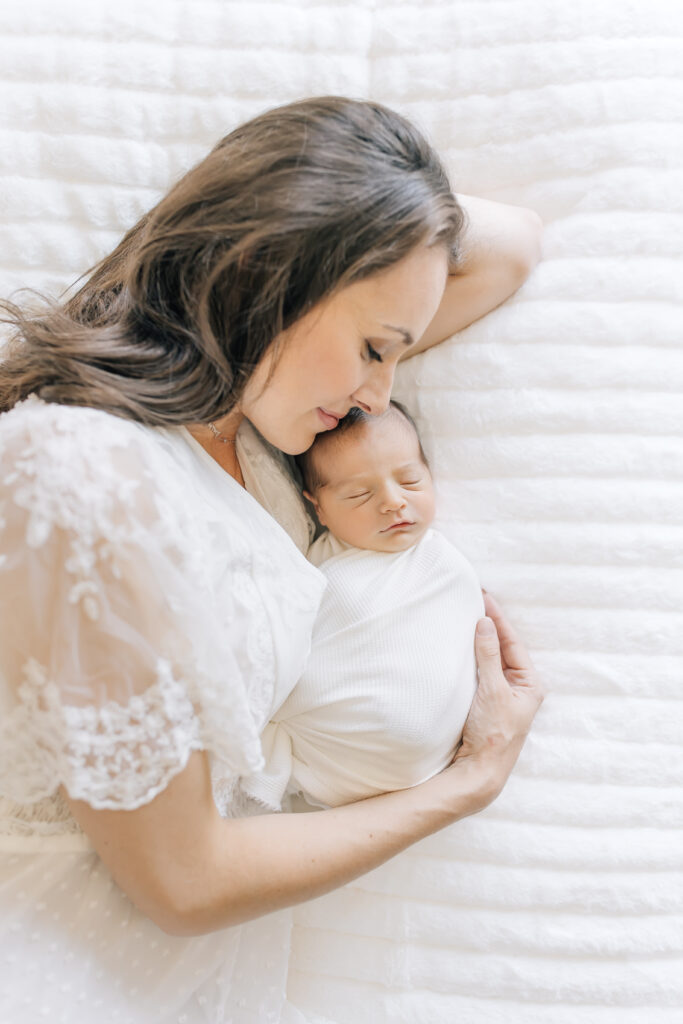 A birds-eye-view mom laying on her left side dressed in a white, lace, flowing short-sleeve maternity gown (left) wraps her arm around her swaddled newborn both laying on a bed with white bedding.


