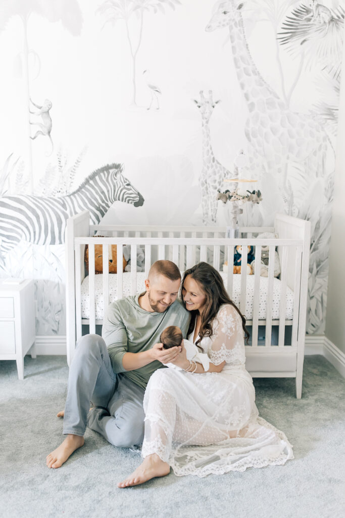A brunette new mom dressed in a white, lace, flowing short-sleeve maternity gown (right) helps hold her swaddled newborn with her husband they sit together on the floor of a grey and white jungle themed nursery in front of the white crib for their in-home newborn photography session.  
