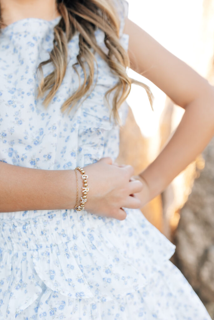 Vertical close up photo with focus on young brunette girls hands on her hip and across her waist wearing a golden lettered bracelet, blue and white floral ruffled sleeveless dress and curled hair. This showcases the magnitude of tiny details in accessories for the perfect spring family photo inspo.
