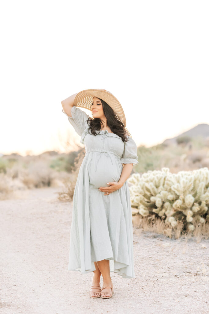A brunette pregnant woman photographed outdoors in the beautiful neutrals of Arizona sand and Chihuahuan Desert wearing a flowing soft teal, blue dress and wide brimmed natural sun hat with one hand around her belly and one hand on her hat for this maternity photoshoot idea for 2024.