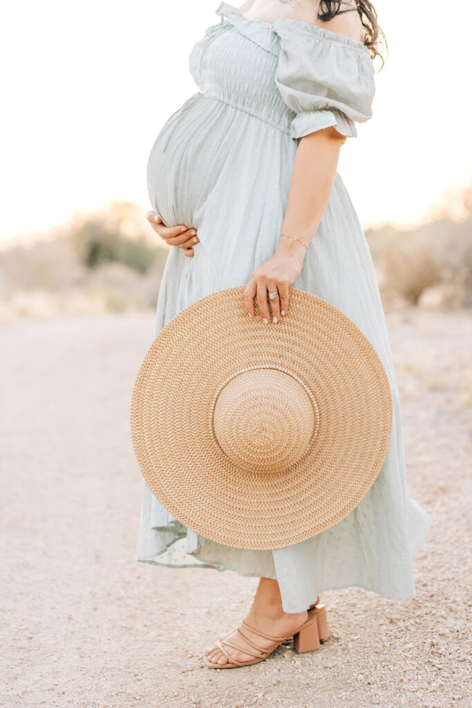 A brunette pregnant woman photographed outdoors in the beautiful neutrals of Arizona sand and Chihuahuan Desert wearing a flowing soft teal, blue dress and wide brimmed natural sun hat with one hand around her belly and one hand holding her hat down for this maternity photoshoot idea for 2024