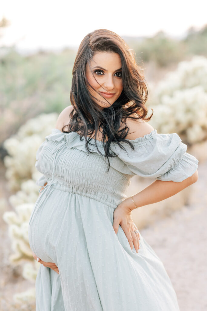 A brunette pregnant woman photographed outdoors in the beautiful neutrals of Arizona sand and Chihuahuan Desert wearing a flowing soft teal, blue dress and looking directly at the camera.