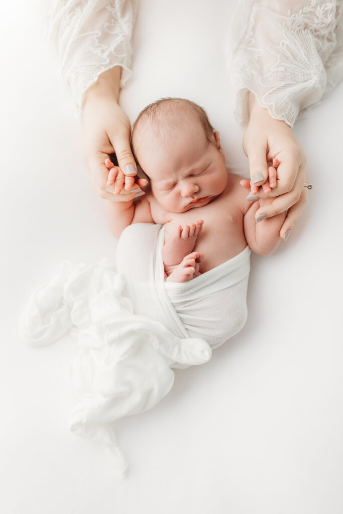 A birds-eye-view of newborn baby with a swaddled bottom laying on a white bedsheet and hands holding moms showcasing a nude/grey nail polish and white, translucent, floral laced long-sleeves for this sweet in-home newborn photography session. 