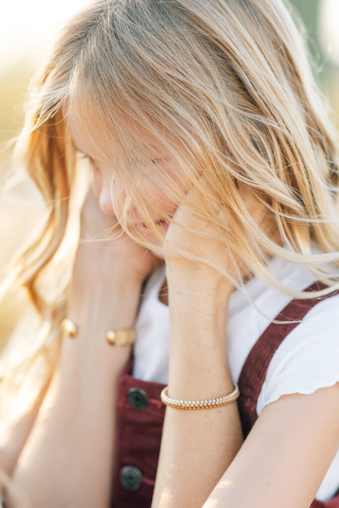 Vertical close up photo with focus on young blonde girls face wearing a white tshirt and maroon overall style stop showing, hair blowing and mom holding her cheeks wearing a gold textured bracelet on one wrist and a solid gold wider bracelet on the other. 
