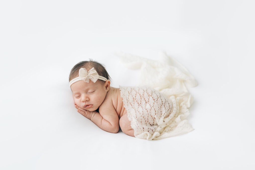 A full body view of newborn baby's side as she is curled up on her belly with arms and legs tucked underneath, head adorned with a neutral colored headband bow and a swatch of cream lace fabric draped over her bottom. Photographed on a pure white background.