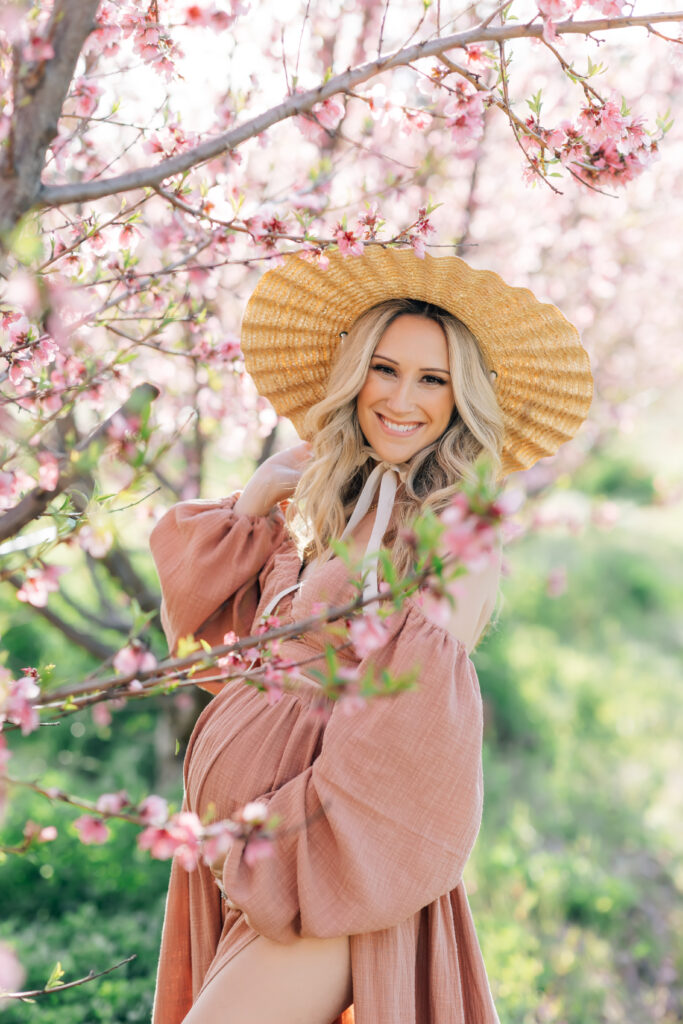 A blonde pregnant woman photographed outdoors surrounded by pink spring blooms wearing a  long-sleeved dusty rose colored dress and wide brimmed natural hat holding her baby bump and looking directly into the camera for a mid-thigh close cropped maternity session image from this maternity photoshoot idea for 2024