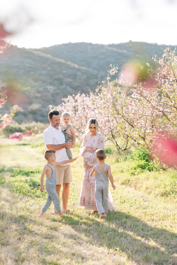Family of 5 interacting together in a field of pink blooms. From left to right, brown haired boy in blue linen overalls, dark haired dad in white collared shirt and khaki shorts holding toddler blonde son in green linen overalls, pregnant mom in long sleeve white and pink floral dress holding above and below her belly with brown haired boy in matching blue overalls for an amazing example of spring family photo outfits.