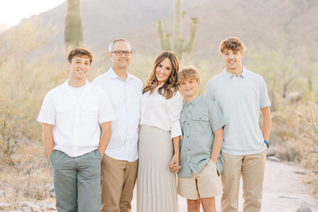 Family of 5 horizontal image cropped at the knees for a classic spring family photo. From left to right, brown curly haired teenage boy in white short sleeve shirt and green pants, dad with white hair and glasses in light blue button up short sleeve top and dark tan pants, brunette mom in white top and neutral colored pleated long skirt, young boy in green button up short sleeve shirt and light khaki shorts and teenage curly haired boy in light blue polo and mid-toned tan khaki pants. Behind the family is a true Arizona landscape filled with cactus, desert sand and Saguaro.