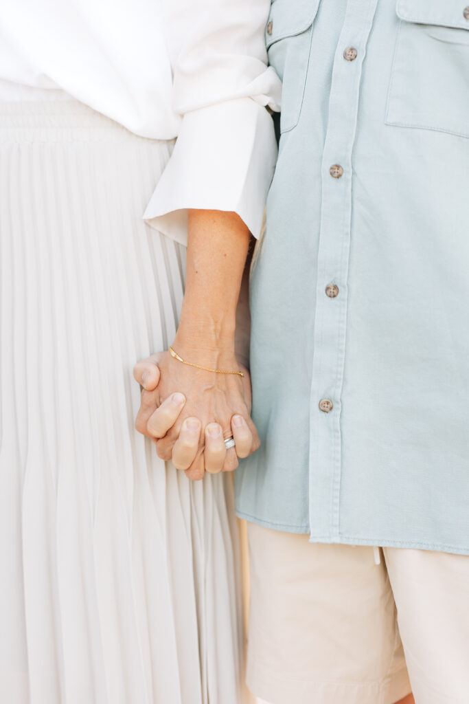 Vertical close up photo highlighting mom and son holding hands with mom wearing a gold bracelet and pearl ring, white top and neutral colored pleated long skirt, young boy in green button up short sleeve shirt and light khaki shorts.