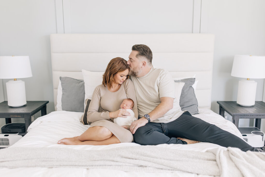 An auburn new mom dressed in a light tan long-sleeve maternity gown (left) wrapped up in and sitting beside a dark haired new dad's arms dressed in a white tee shirt and black jeans (right) holding their swaddled newborn photographed in their master bedroom on the bed. The room is very neutral cool tones with lots of white and grey with two identical black wooden nightstands topped with white lamps on either side of the bed. 