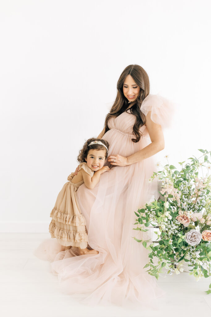 A brunette pregnant woman dressed in a peachy pink fluffy maternity gown looking down at her belly and daughter snuggled up to her wearing a neutral tan colored ruffled dress photographed on a studio white backdrop with stunning fresh florals