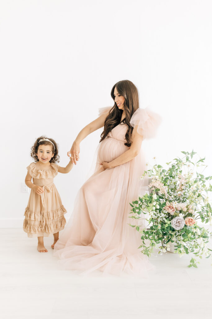 A brunette pregnant woman dressed in a peachy pink fluffy maternity gown twirling her daughter wearing a neutral tan colored ruffled dress photographed on a studio white backdrop with stunning fresh florals