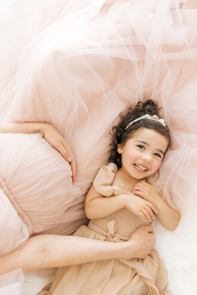 A close up vertical image with the focus on a pregnant woman's belly dressed in a peachy pink fluffy maternity gown and her daughter snuggled up to her wearing a neutral tan colored ruffled dress smiling and looking at the camera photographed on a studio white backdrop and from an arial point of view