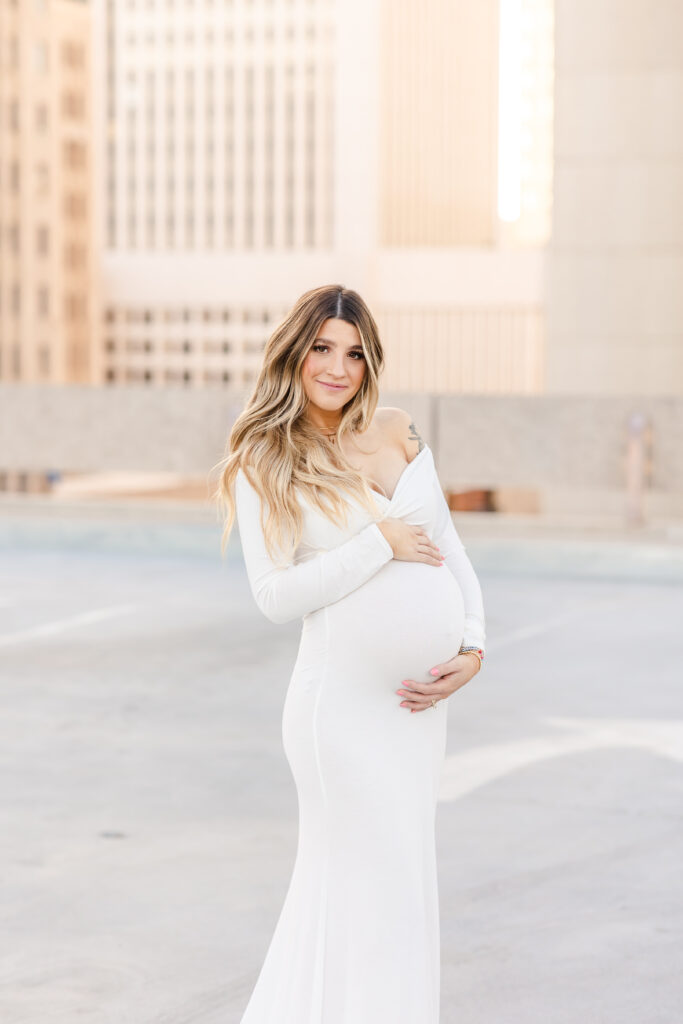 A dirty blonde pregnant woman photographed outdoors on a rooftop wearing a stunning white off the shoulder sweetheart neckline long-sleeved dress holding her baby bump with both hands and looking directly into the camera for this maternity photoshoot idea for 2024.