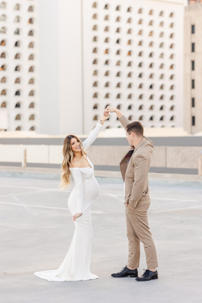 A dirty blonde pregnant woman photographed outdoors on a rooftop wearing a stunning white off the shoulder sweatheart neckline long-sleeved dress being twirled around by her husband wearing a tailored tan suit and black dress shoes. 