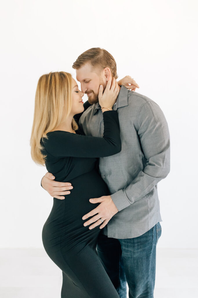 A blonde pregnant woman dressed in a black long-sleeve maternity gown (left) wrapped up in a brown haired guys arms dressed in a grey long sleeve button up shirt and jeans (right) photographed on a studio white backdrop 