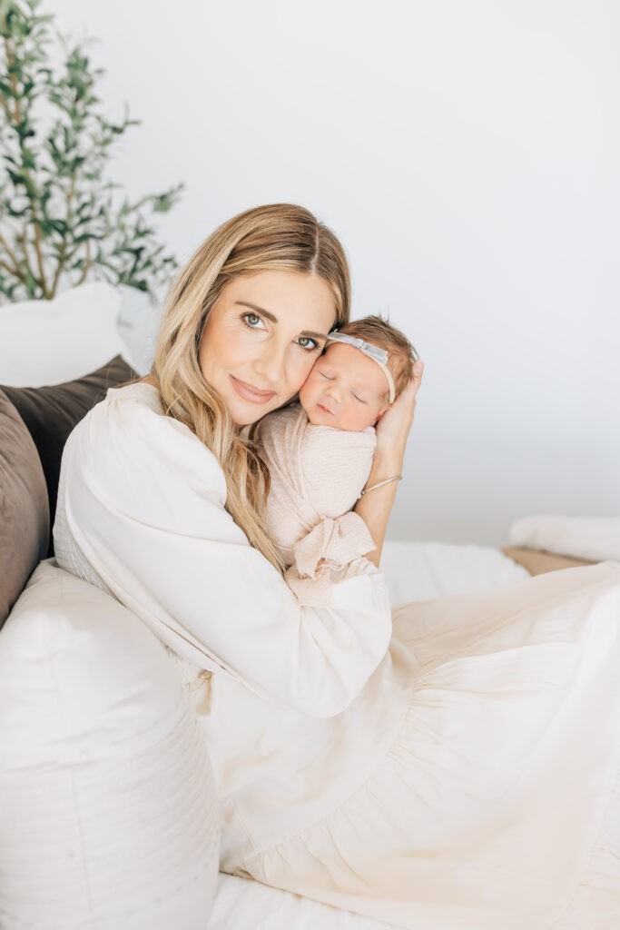 A blonde mom with long hair holding newborn baby girl swaddled in sand colored swaddle and sand headband with soft blue colored bow. Mom is sitting sideways on a terracotta bed and white pillows in an all white room and small bit of olive tree branches peeking in from camera left. 