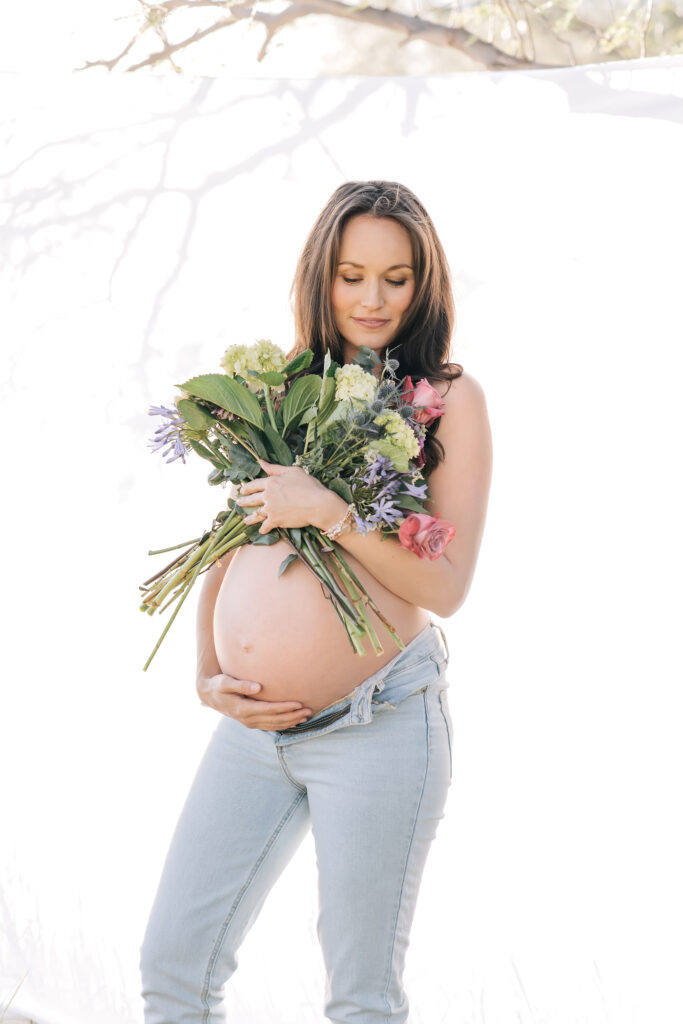 A brunette pregnant woman photographed outdoors in a field with a white sheet hung up in the trees. She is holding her bare belly in one hand and a bright and colorful bouquet in the other wearing light-wash jeans and looking down at the fresh bouquet in this closer up image of this amazing maternity photoshoot idea