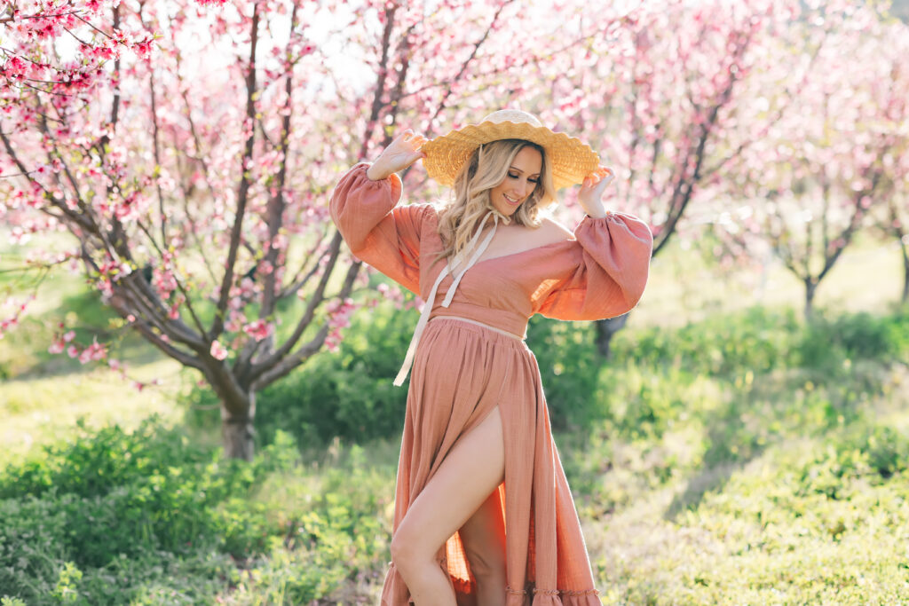 A blonde pregnant woman photographed outdoors surrounded by pink spring blooms wearing a  long-sleeved dusty rose colored dress and holding her wide brimmed natural hat, looking down and to camera right.