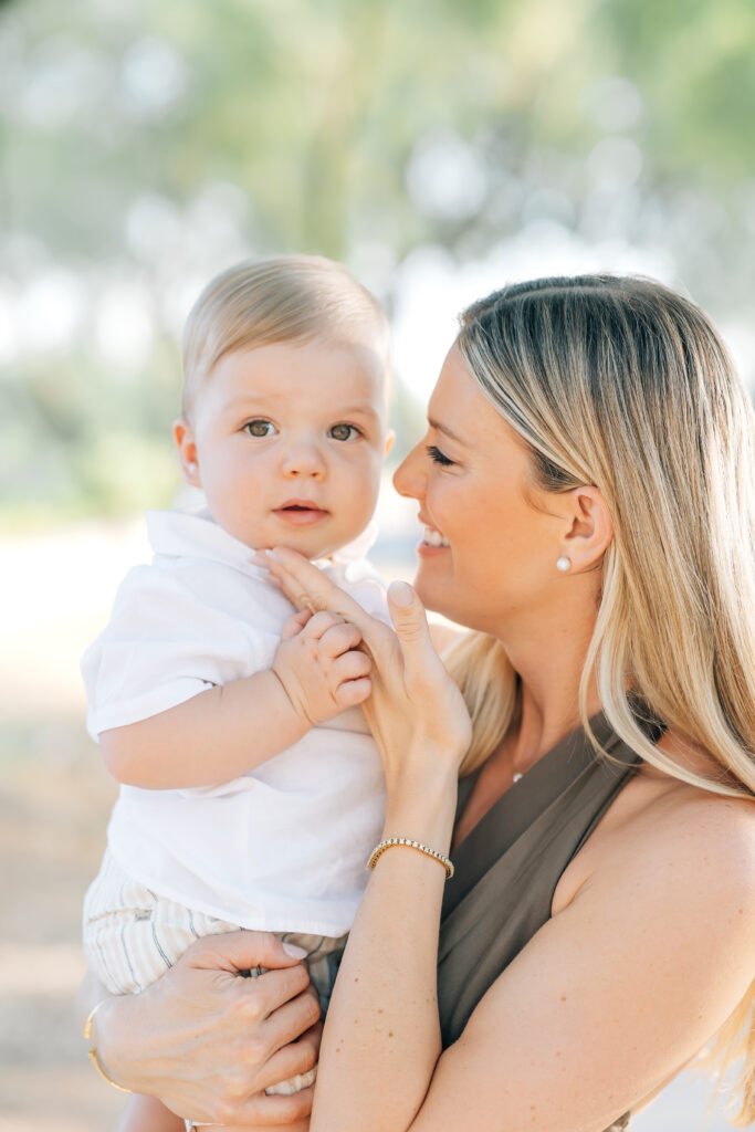 Close-up vertical photo of mom and toddler boy surrounded by light Arizona earthy tones of trees. Right, blonde mom smiling at son and touching his chin wearing a satin, brown maxi halter dress. pearl stud earrings and diamond tennis bracelet holding toddler son wearing a white button-up shirt, khaki shorts looking at the camera and touching mom's hand. 

