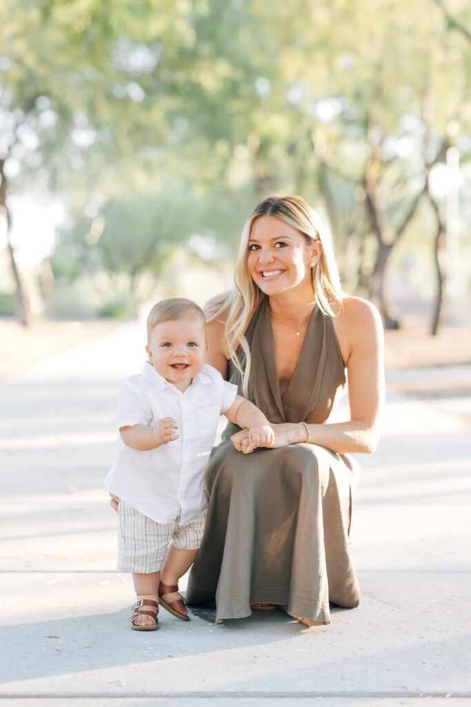Full body vertical photo of mom and toddler boy surrounded by light Arizona earthy tones of trees and ground. Mom crouched down on sidewalk as son stands and uses her legs/arm for support. Right, blonde mom smiling at camera and wearing a satin, brown maxi halter dress. pearl stud earrings, necklace and diamond tennis bracelet arm around toddler son wearing a white button-up shirt, khaki shorts looking at the camera. 