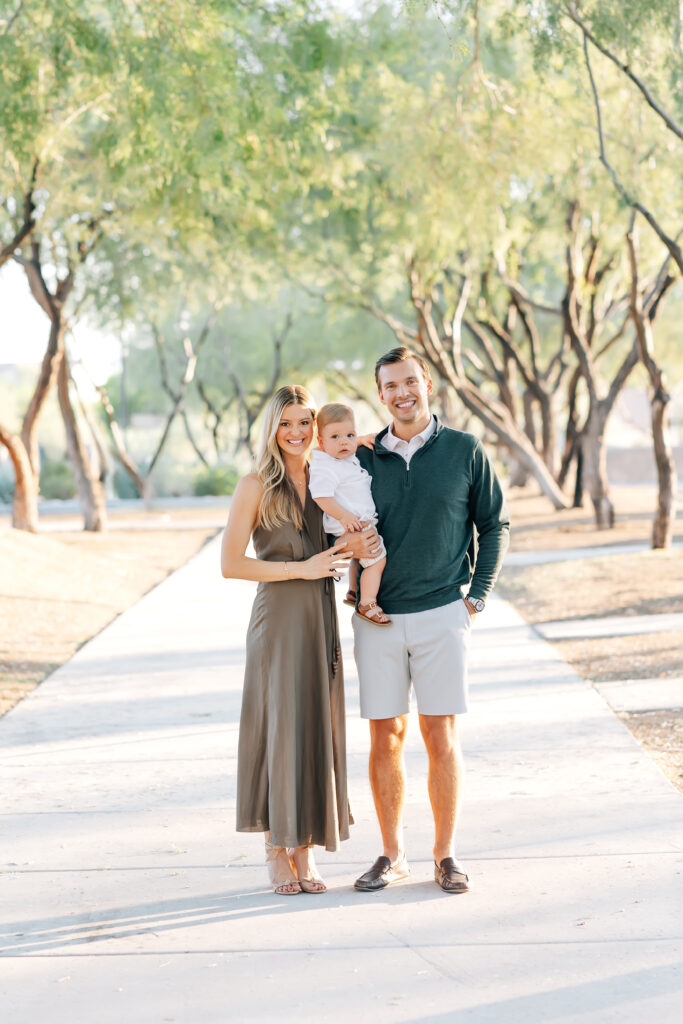 Full length vertical outdoor photo of a family of 3 standing on a white sidewalk surrounded by light Arizona earthy tones of trees and ground for their mini-session photos. Left, blonde mom wearing a satin, brown maxi halter dress helping to hold dirty blonde toddler son dressed in a white button-up shirt, khaki shorts and brown leather sandals. Dark brunette Husband, camera right, holding toddler son wearing a white button up collared shirt underneath an emerald green half-zip sweater and light grey tailored shorts. Entire family looking and smiling at the camera.