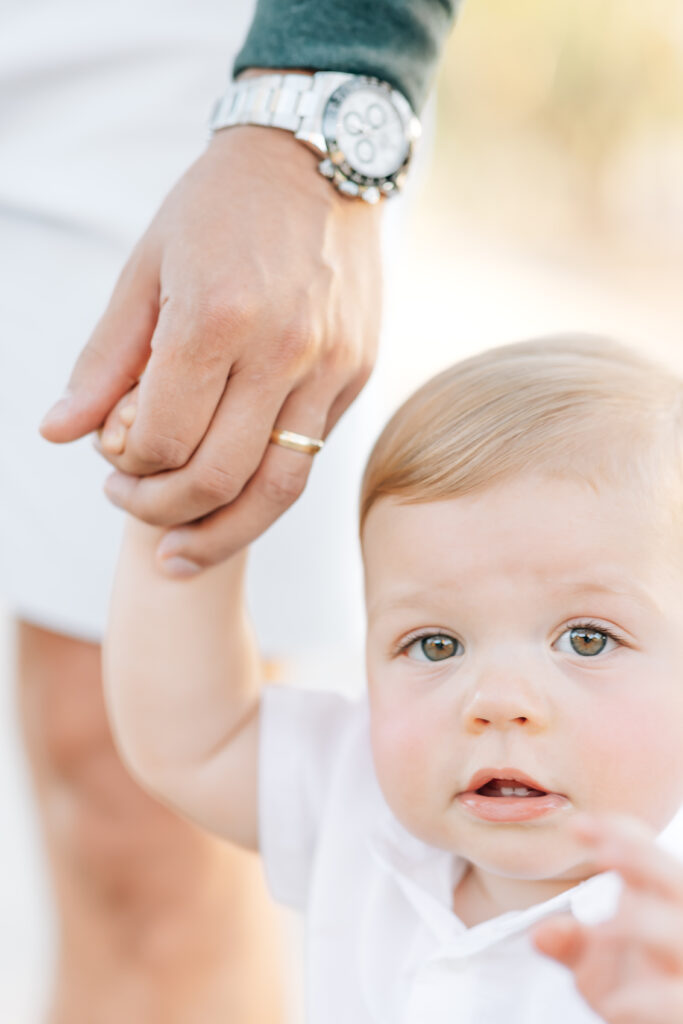 Close-up vertical photo of toddler boy holding the hand of dad showcasing gold wedding band and silver watch peaking out of his emerald green sleeve. Toddler son wearing a white button-up shirt, reaching out toward the camera.
