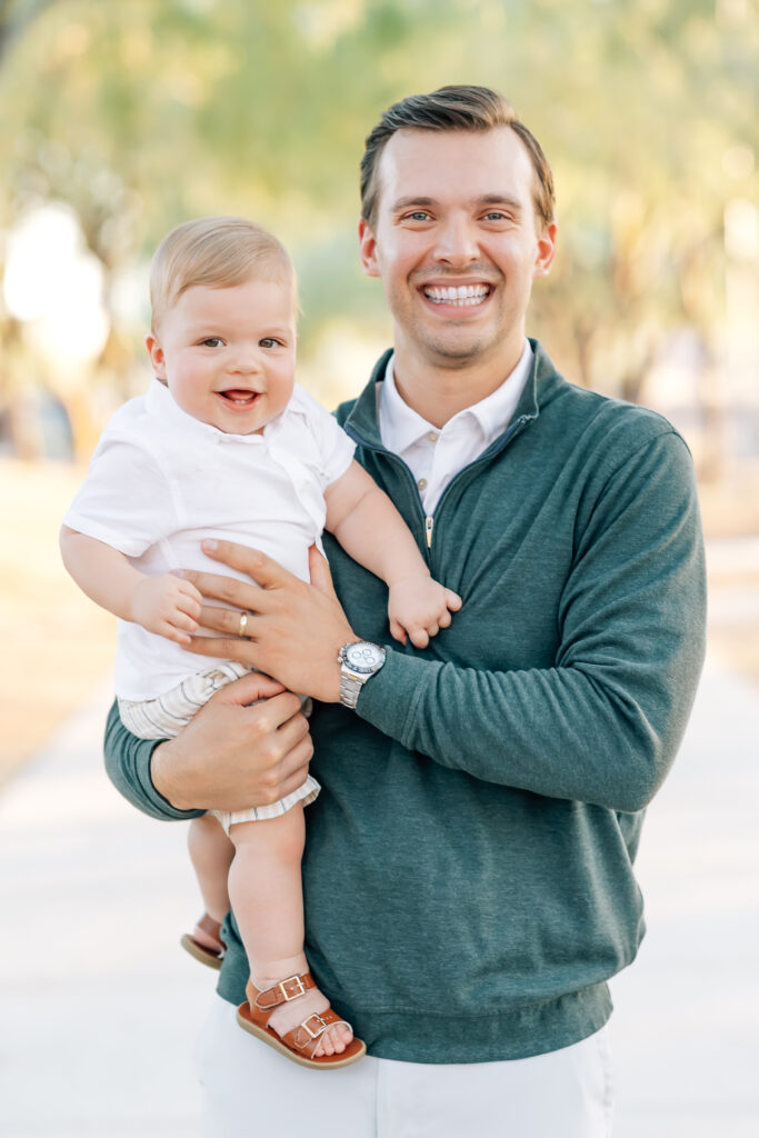 Close-up vertical photo of dad and toddler boy surrounded by light Arizona earthy tones of trees. Right, blonde Dark brunette dad, camera right, wearing a white button up collared shirt underneath an emerald green half-zip sweater and light grey tailored shorts holding toddler son wearing a white button-up shirt, khaki shorts looking at the camera and smiling. 
