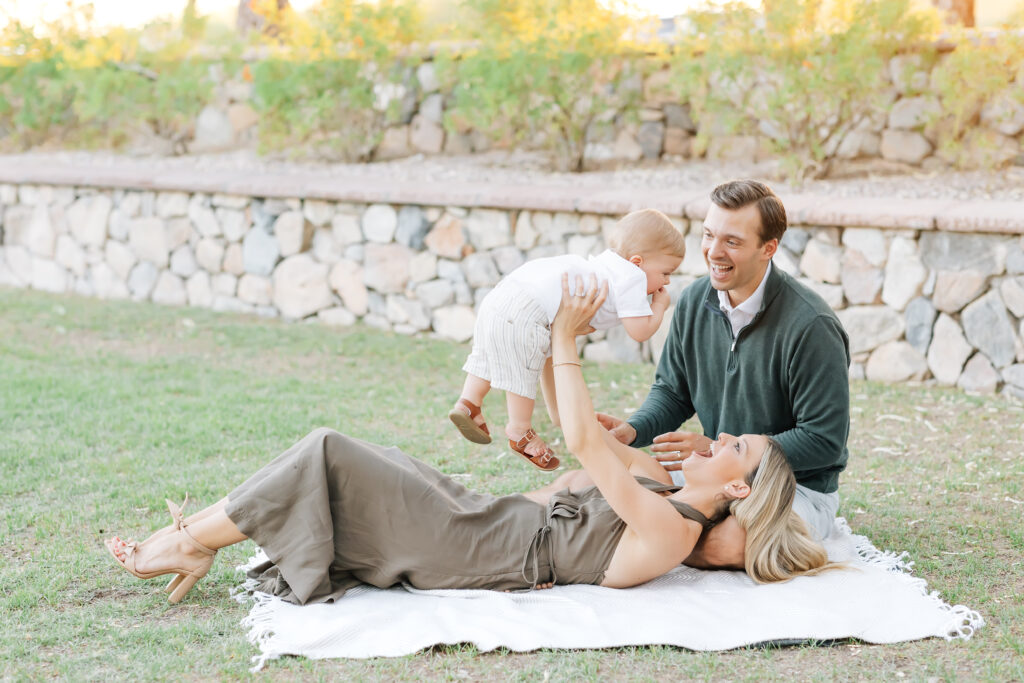 Horizontal full body photo of a family of 3 in the grass on a blanket. Mom holding toddler son in the air while laying on her back with husband sitting up laughing and smiling at their son for their mini- session photos. Blonde mom smiling at son wearing a satin, brown maxi halter dress. Dark brunette Husband, camera right, wearing a white button up collared shirt underneath an emerald green half-zip sweater and light grey tailored shorts. Dirty blonde, toddler boy dressed in a white button-up shirt, khaki shorts and brown leather sandals. 
