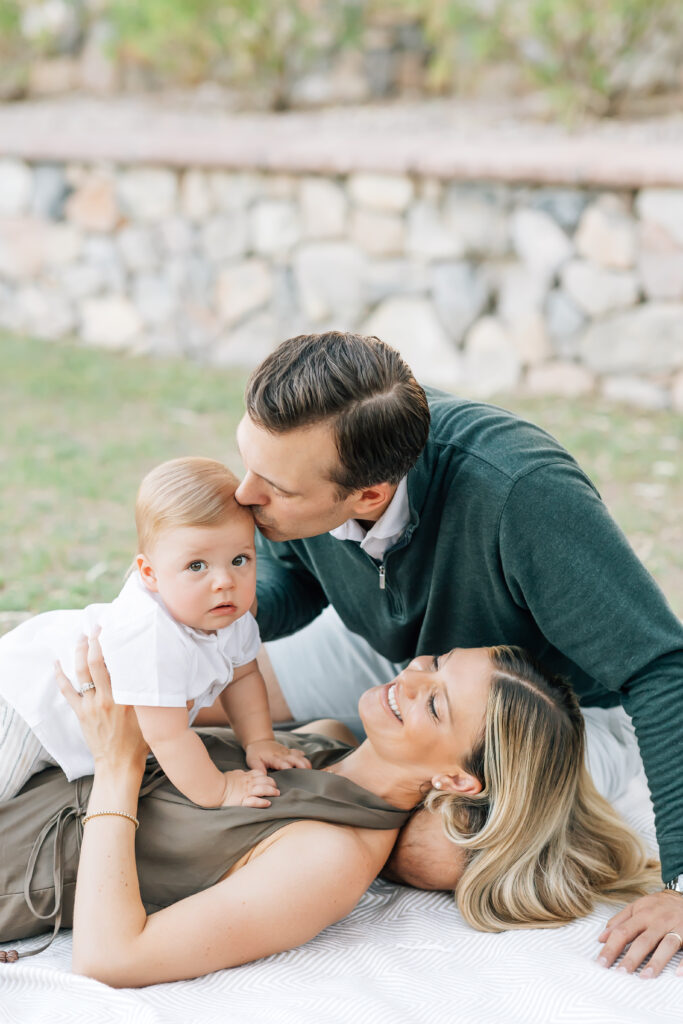 Vertical mid-crop photo of a family of 3 in the grass on a blanket. Mom holding toddler son on her stomach while laying on her back with husband sitting up and leaning over to kiss their son for their mini session photoshoot. Blonde mom smiling at son wearing a satin, brown maxi halter dress. Dark brunette Husband, camera right, wearing a white button up collared shirt underneath an emerald green half-zip sweater and light grey tailored shorts. Dirty blonde, toddler boy dressed in a white button-up shirt and khaki shorts. 