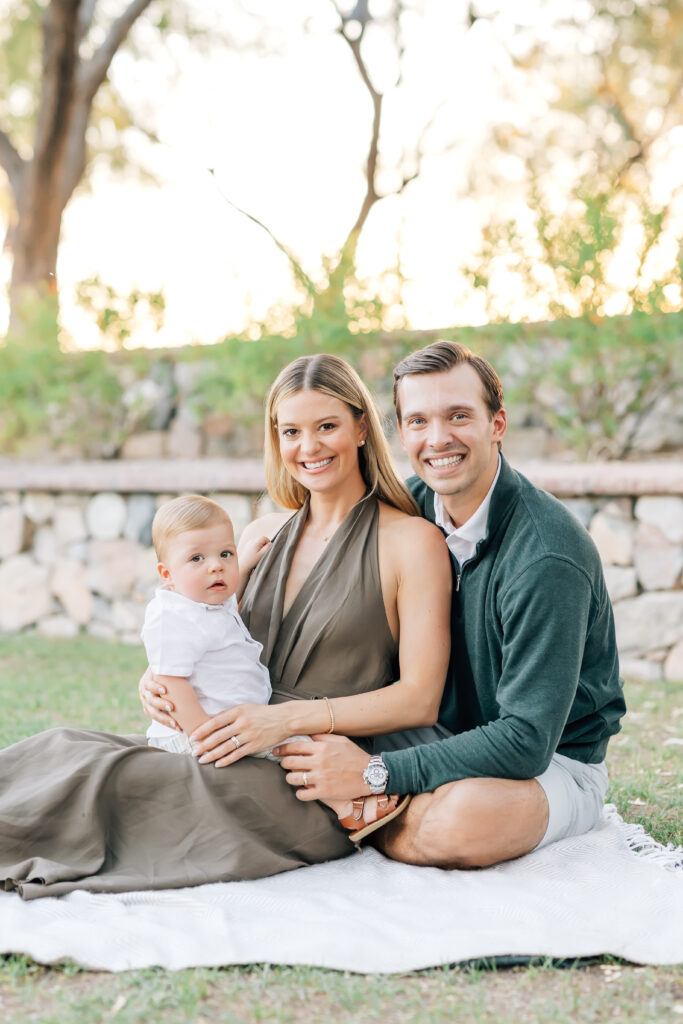 Mid-crop vertical photo of a family of 3 sitting on the ground with a light grey blanket all looking toward the camera for their mini session photoshoot. Center, blonde mom wearing a satin, brown maxi halter dress holding toddler son on her lap. Dark brunette Husband, camera right, arms wrapped around his wife wearing a white button up collared shirt underneath an emerald green half-zip sweater and light grey tailored shorts. Dirty blonde toddler son dressed in a white button-up shirt, khaki shorts and brown leather sandals sitting facing mom belly-to-belly. 
