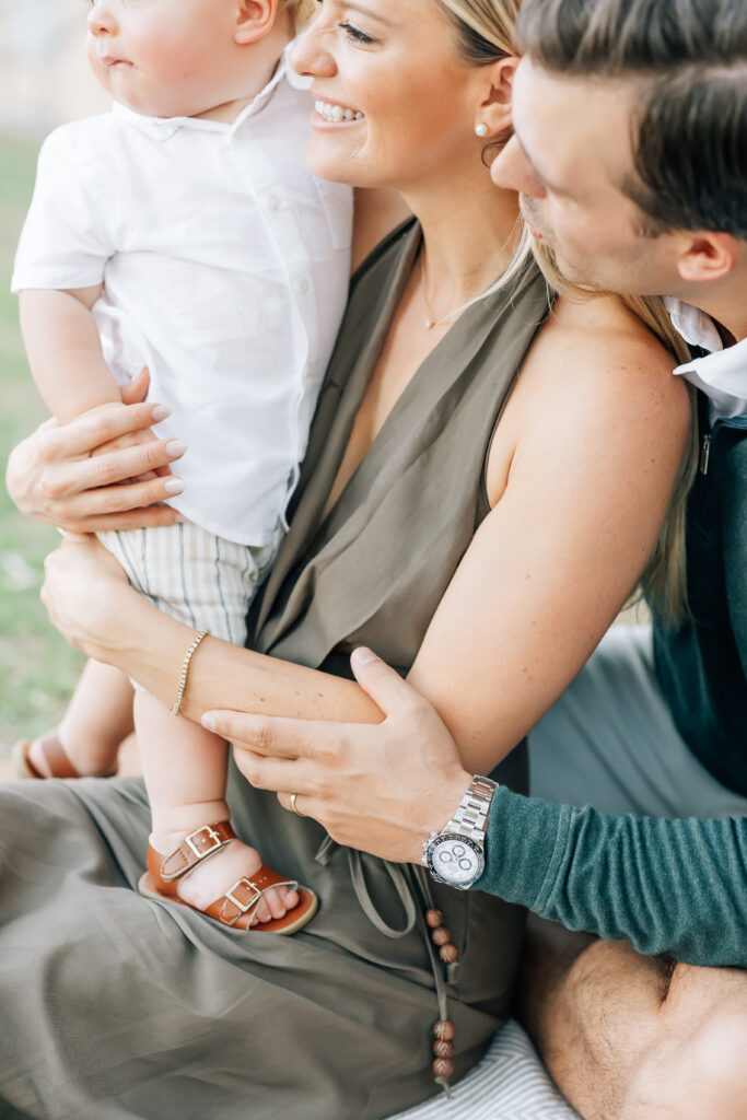 Vertical close-up photo of a family of 3 sitting in the grass on a blanket looking off camera left. Mom holding toddler son standing on her lap with husband sitting up and leaning over her shoulder. Blonde mom wearing a satin, brown maxi halter dress. Dark brunette Husband, camera right, wearing a white button up collared shirt underneath an emerald green half-zip sweater and light grey tailored shorts. Dirty blonde, toddler boy dressed in a white button-up shirt and khaki shorts. Detail mini-session photos like this make for the best memories!
