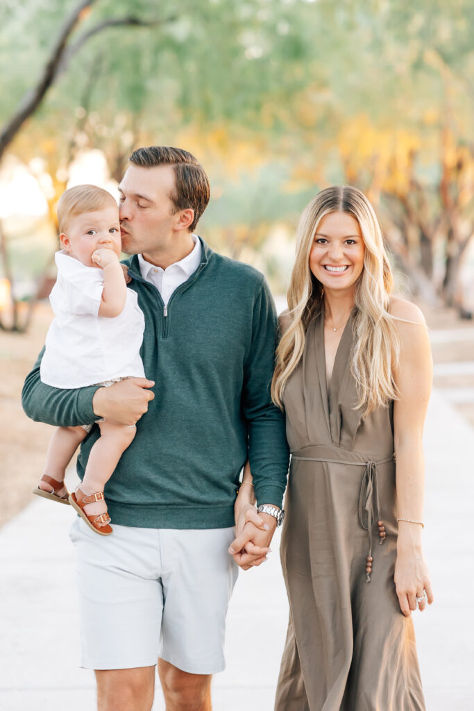 Mid-crop vertical photo of a family of 3 walking toward the camera on a white sidewalk surrounded by light Arizona earthy tones of trees and ground for their mini session photoshoot. Right, blonde mom smiling at the camera wearing a satin, brown maxi halter dress holding hands with her husband. Dark brunette Husband, camera right, holding toddler son wearing a white button up collared shirt underneath an emerald green half-zip sweater and light grey tailored shorts. Holding and kissing the cheek of dirty blonde toddler son dressed in a white button-up shirt, khaki shorts and brown leather sandals. 
