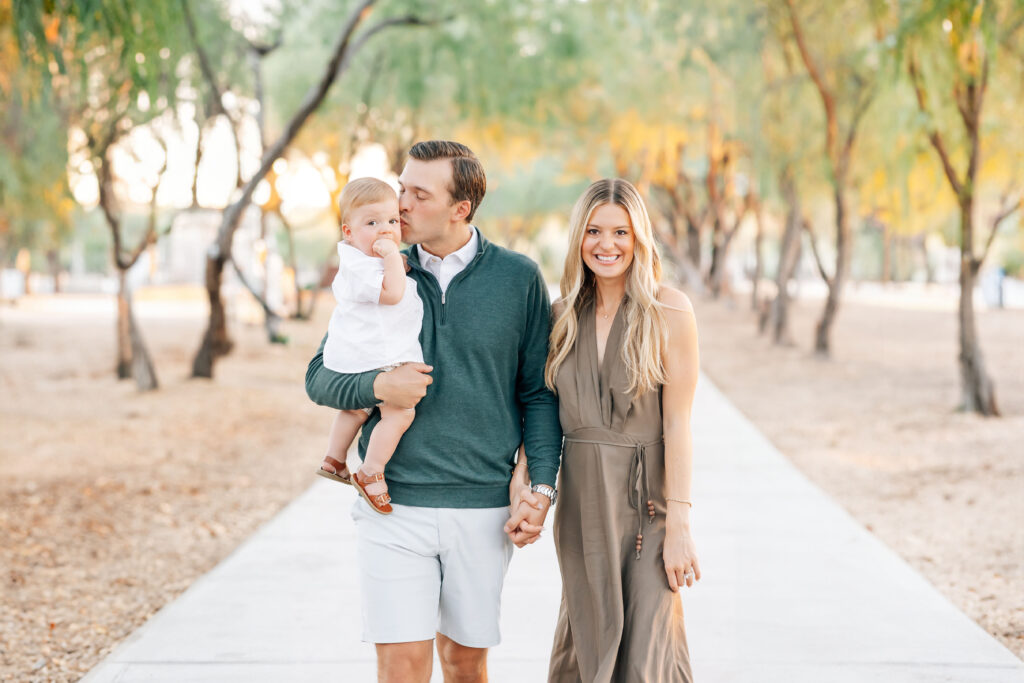 Mid-crop horizontal photo of a family of 3 walking toward the camera on a white sidewalk surrounded by light Arizona earthy tones of trees and ground for their mini session photoshoot. Right, blonde mom smiling at the camera wearing a satin, brown maxi halter dress holding hands with her husband. Dark brunette Husband, camera right, holding toddler son wearing a white button up collared shirt underneath an emerald green half-zip sweater and light grey tailored shorts. Holding and kissing the cheek of dirty blonde toddler son dressed in a white button-up shirt, khaki shorts and brown leather sandals. 
