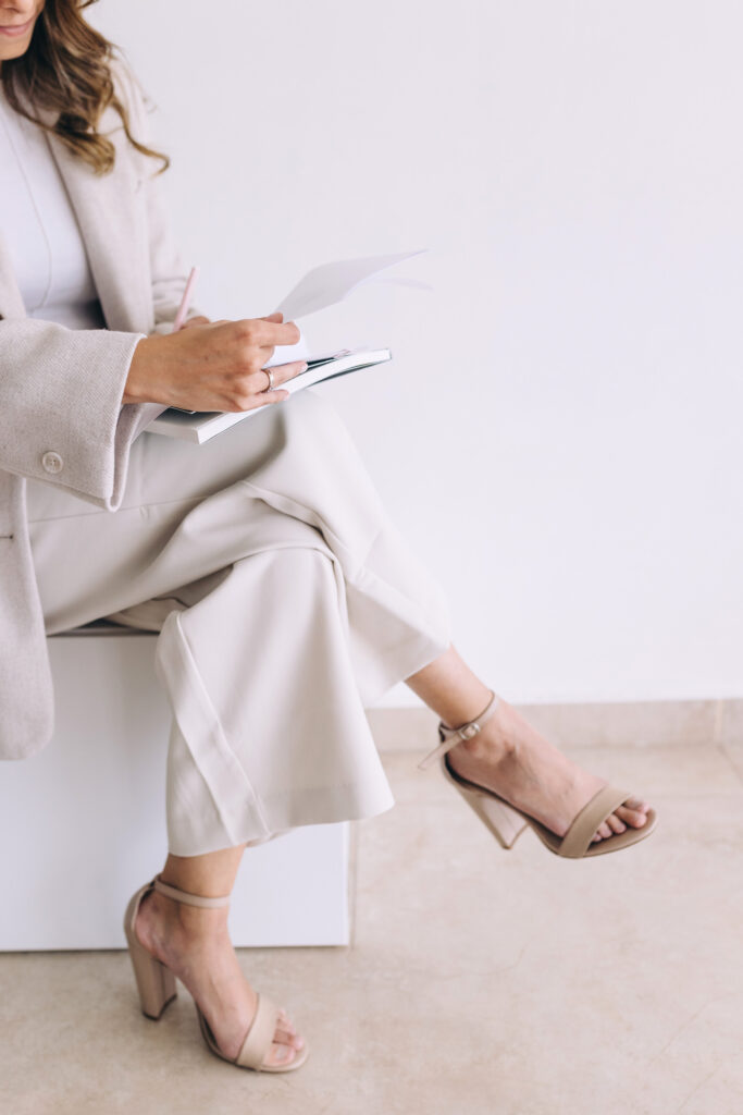 zvertical photo capturing a woman in a beige pant suit with her legs crossed and a notebook on her lap. She is holding a pink pen and looking over papers. The perspective is only of half her body, as the rest is cut of by the composition of the photo. 