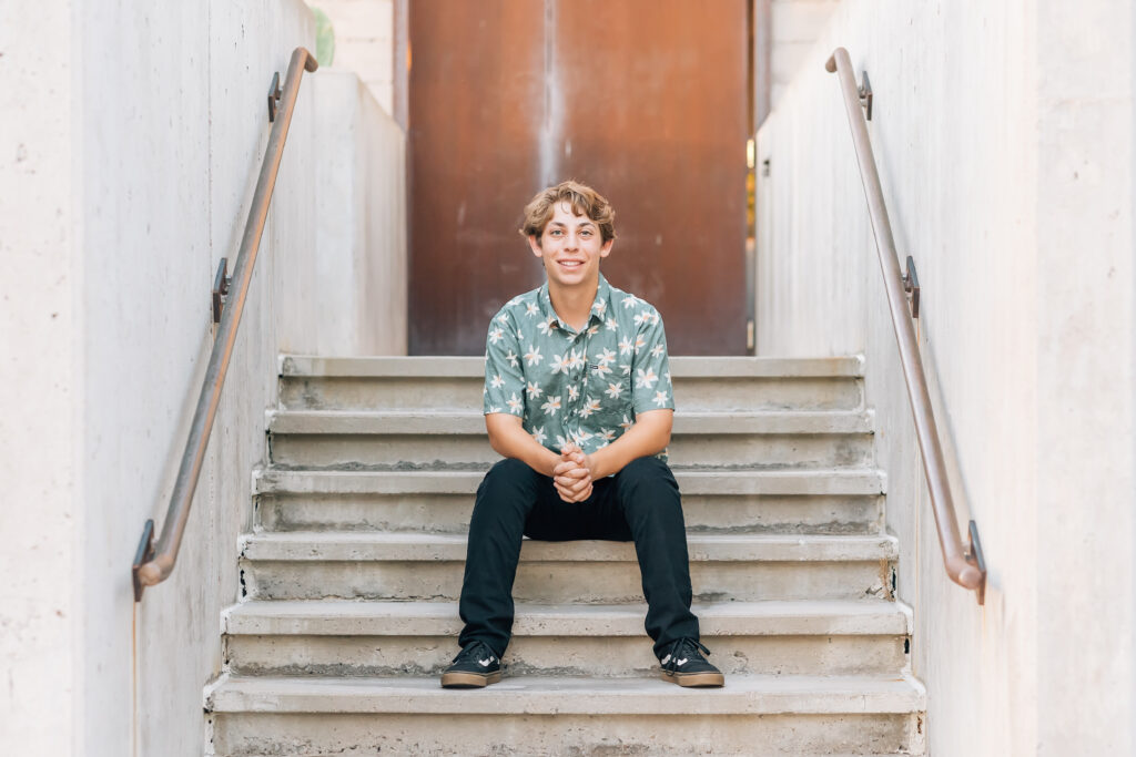 Scottsdale high School senior in black pants and green Hawaiian button up shirt sitting on concrete steps smiling at the camera. 