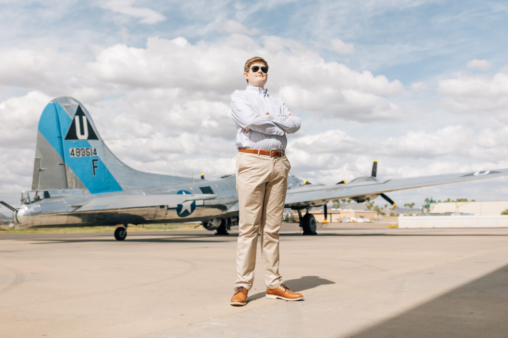 Mountain View Mesa, AZ high school senior wearing a blue button up shirt, khaki pants, and and brown belt with his arms crossed and aviators on while standing in front of  a U.S. Air Force plane. 