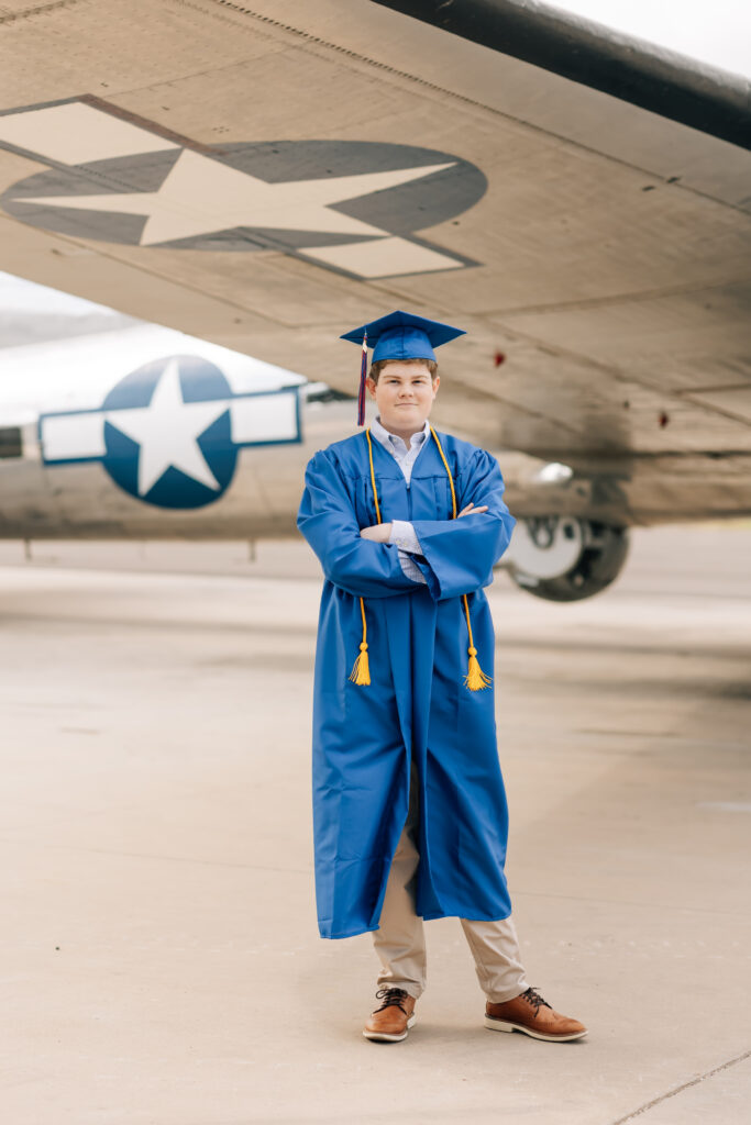 Mountain View Mesa, AZ high school senior wearing his blue cap and gown with a gold cord over his shoulders standing underneath the wing of a U.S. Air Force plane. 